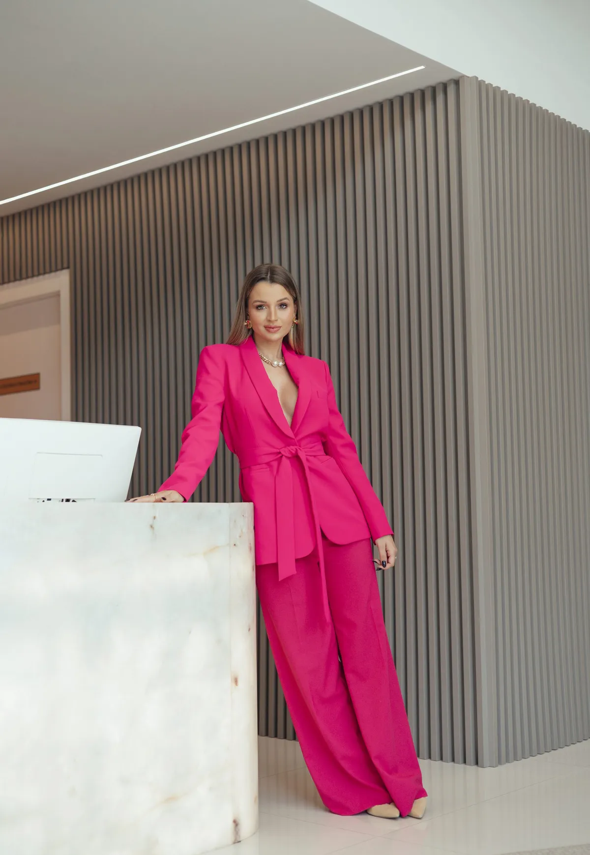 Pricila Hermes, fundadora da La Primore, de terninho pink no balcão de ônix da clínica, diante da parede ripada